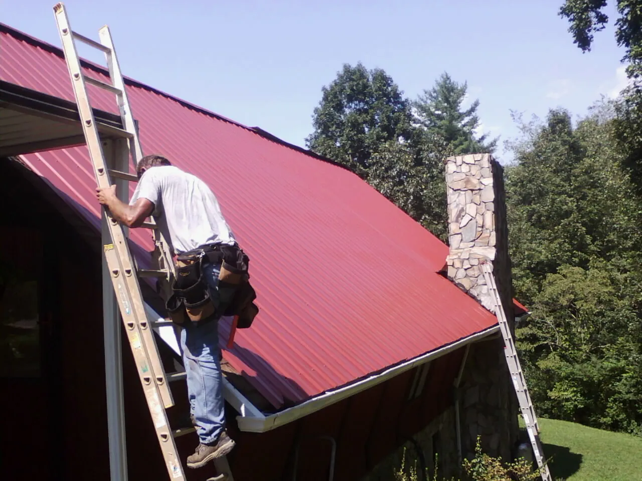 Roofer on a ladder beside a red metal roof and stone chimney — Soffit & Fascia Repair by Roofing Brothers in Scandinavia